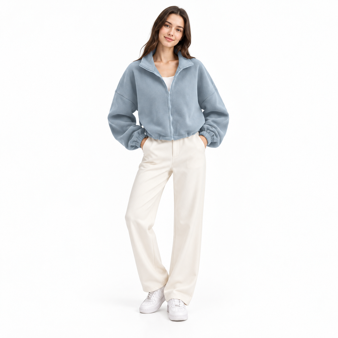 Light gray blue cropped jacket on a full-body model, with an open stand collar, white top, ivory trousers and white backdrop.