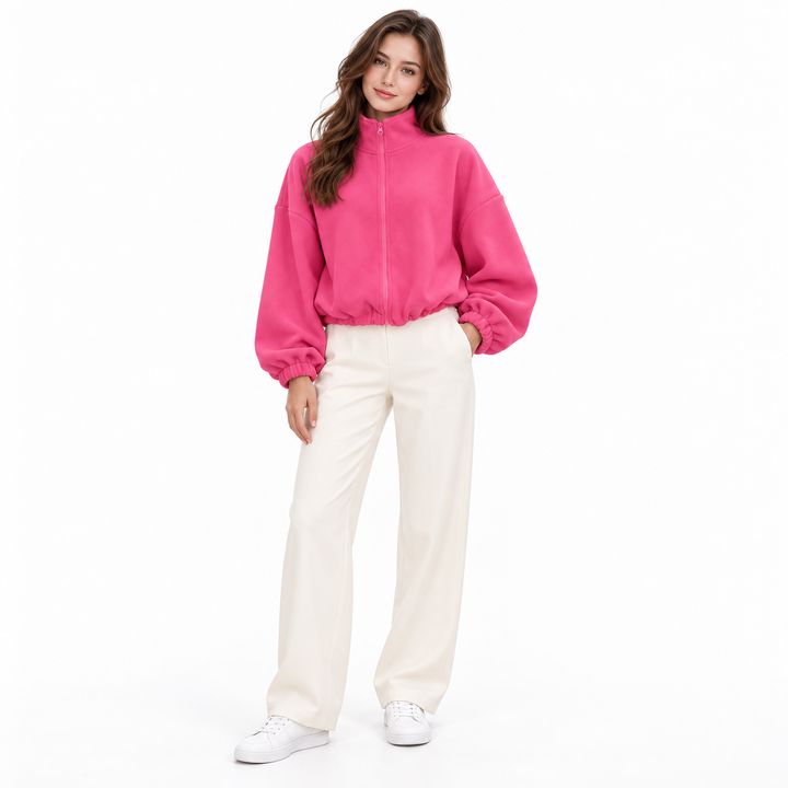 Rose pink cropped jacket on a full-body brunette model showing its stand collar, zipper and gathered hem with ivory trousers.