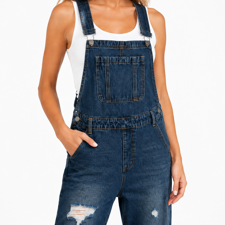 Dark blue cropped denim overalls close-up showing adjustable straps, metal fasteners, bib pocket, seams and distressed denim.