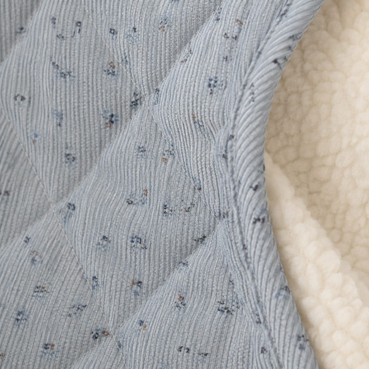 Closeup of blue quilted floral coat texture showing fine ribbed surface, diagonal stitching, printed detail and cream lining.