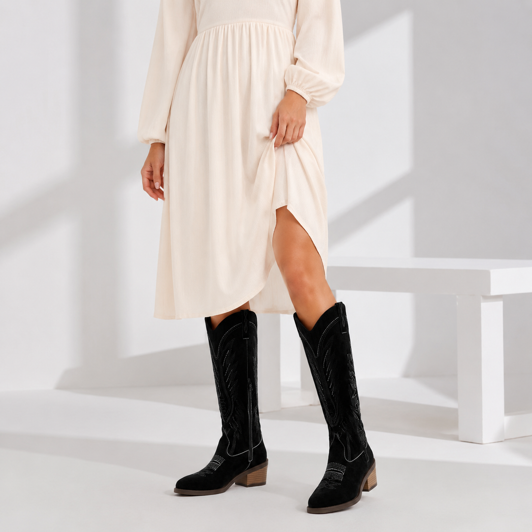 Adult woman styling Black V-cut suede boots in a cream dress, showing embroidered knee-high shafts and stacked heels clearly.