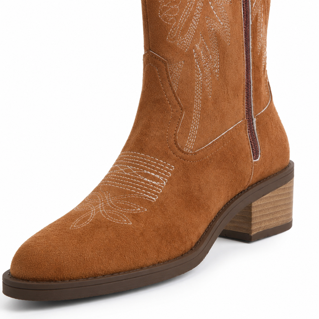 Brown suede boot detail showing stitched toe design, side zipper, sole edge and stacked square heel in a white studio closeup